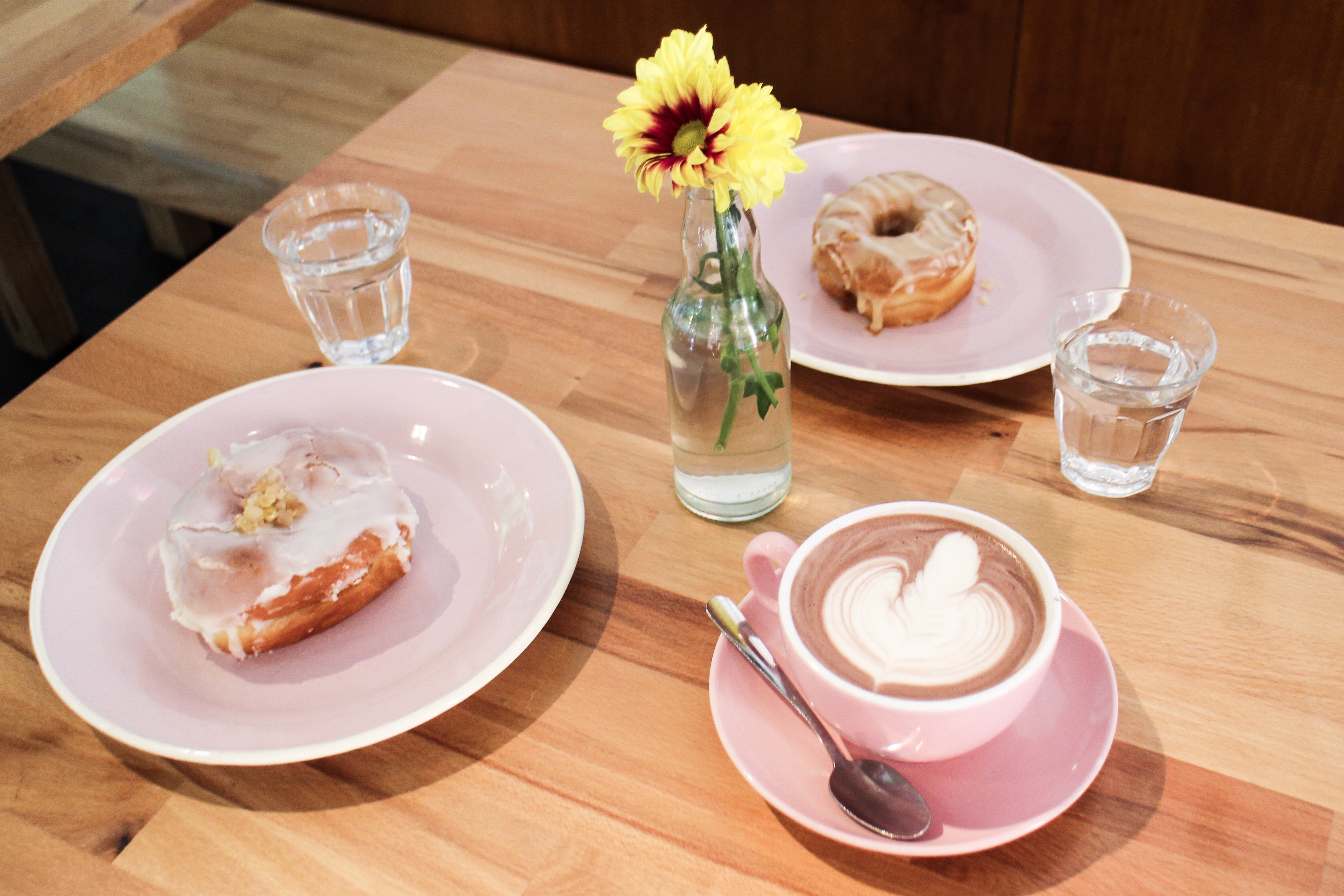 A flatlay consisting of an oat hot chocolate, a G&T doughnut, a lemon & earl grey doughnut and a little vase of flowers. 