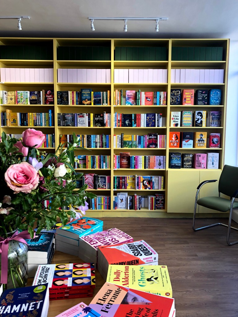 A picture of Rare Birds Book Shop interior in Edinburgh. In the background is a large yellow bookcase that covers an entire wall filled with lots of colourful books. In the foreground is a round display table with a vase of flowers and books facing upwards in a display.