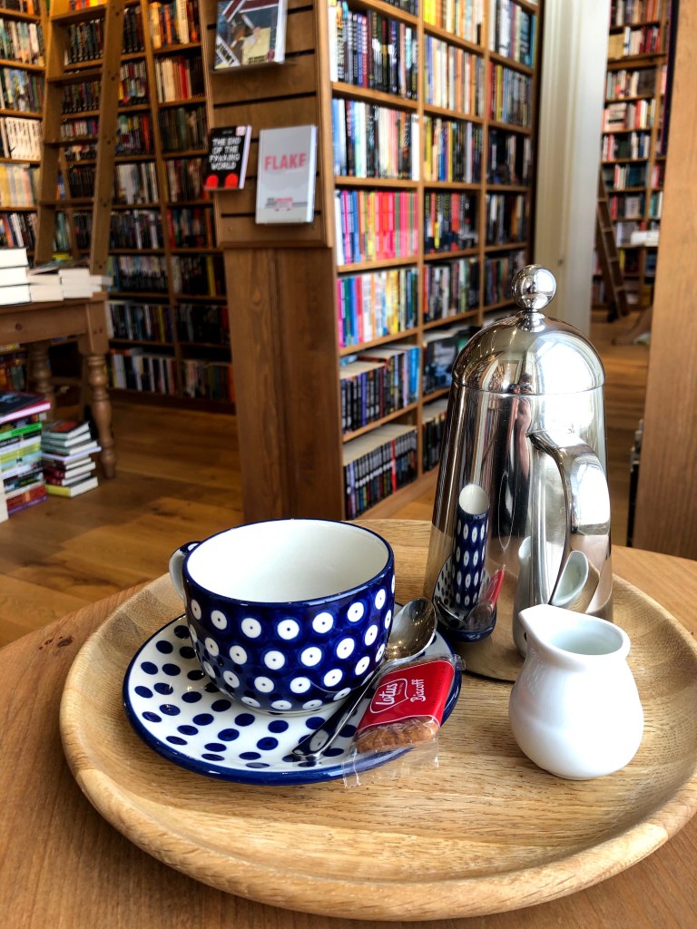 Topping & Company, Edinburgh. a spotty teacup and pot of coffee sit on a small coffee table. It is situated in front of large shelves completely filled with books. 