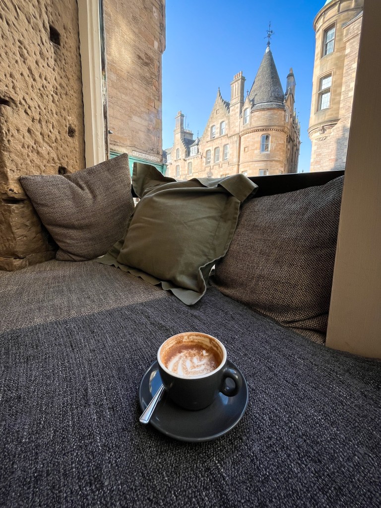 a bench sat in front of a large window. There's muted grey cushions and a blanket giving the window seat a cosy atmosphere and a half drank coffee is perched on top. Through the window are old buildings that are ornately decorated and a blue sky.
