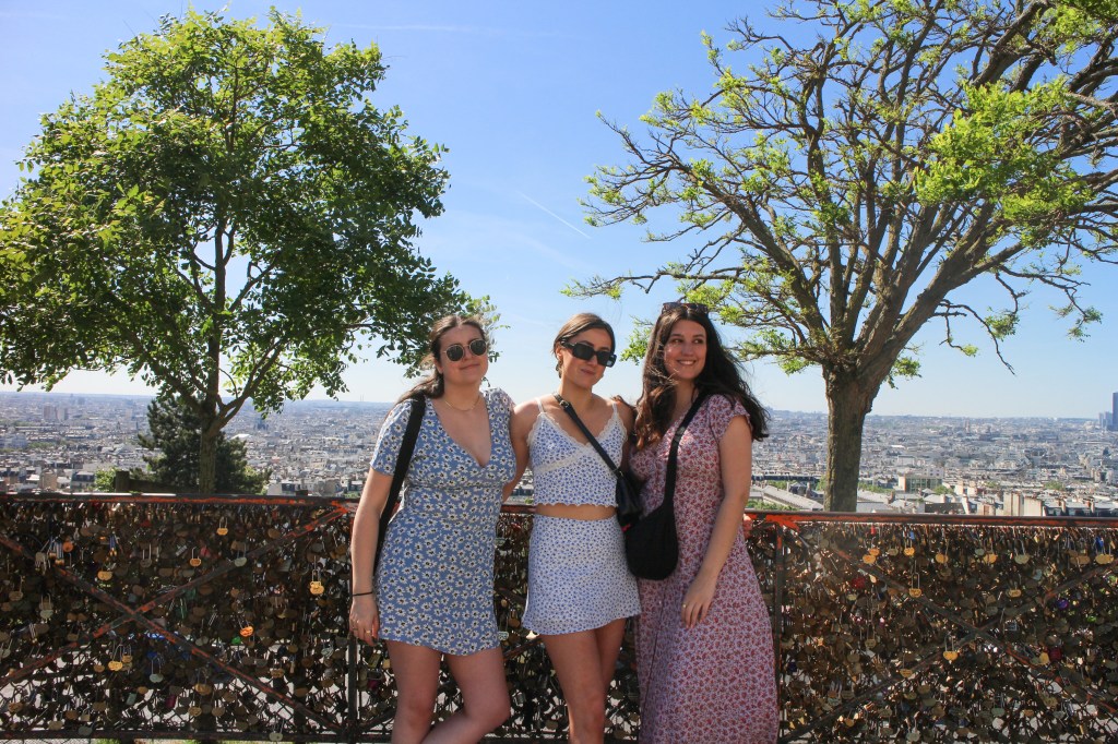 three girls in floral outfits smiling for a photo in front of a railing cover in padlocks. a view of paris is behind them