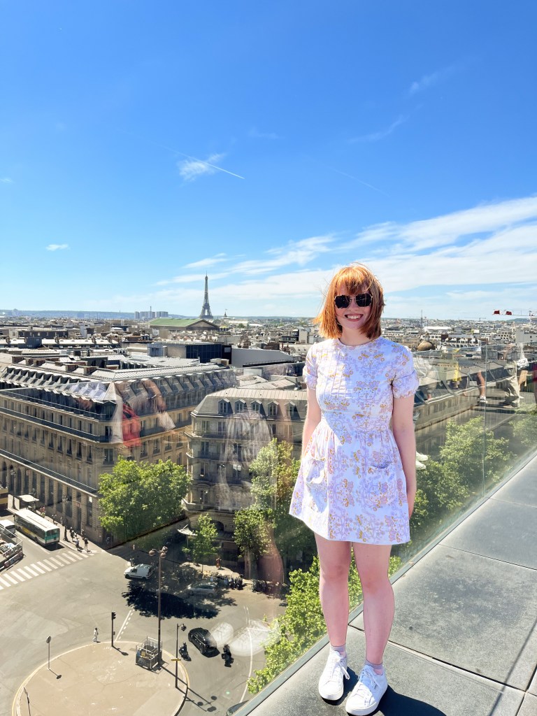 a redheaded gal in a white, floral dress smiling for a photo with a rooftop view of paris