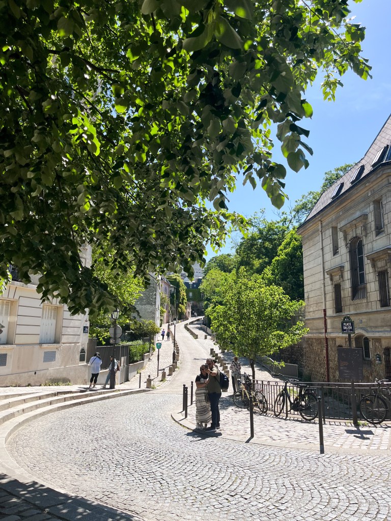 a narrow parisian street that winds up a hill. the road is cobbled and features trees up the pavement