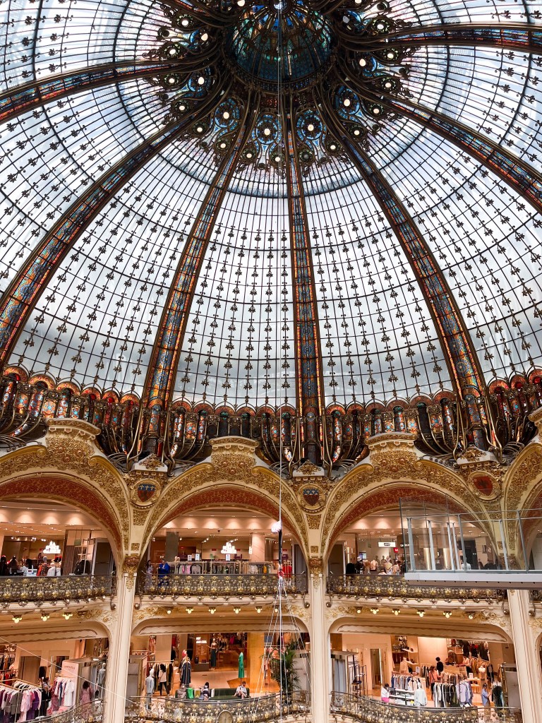an enormous and intricately decorated glass dome above gold arches of a department store