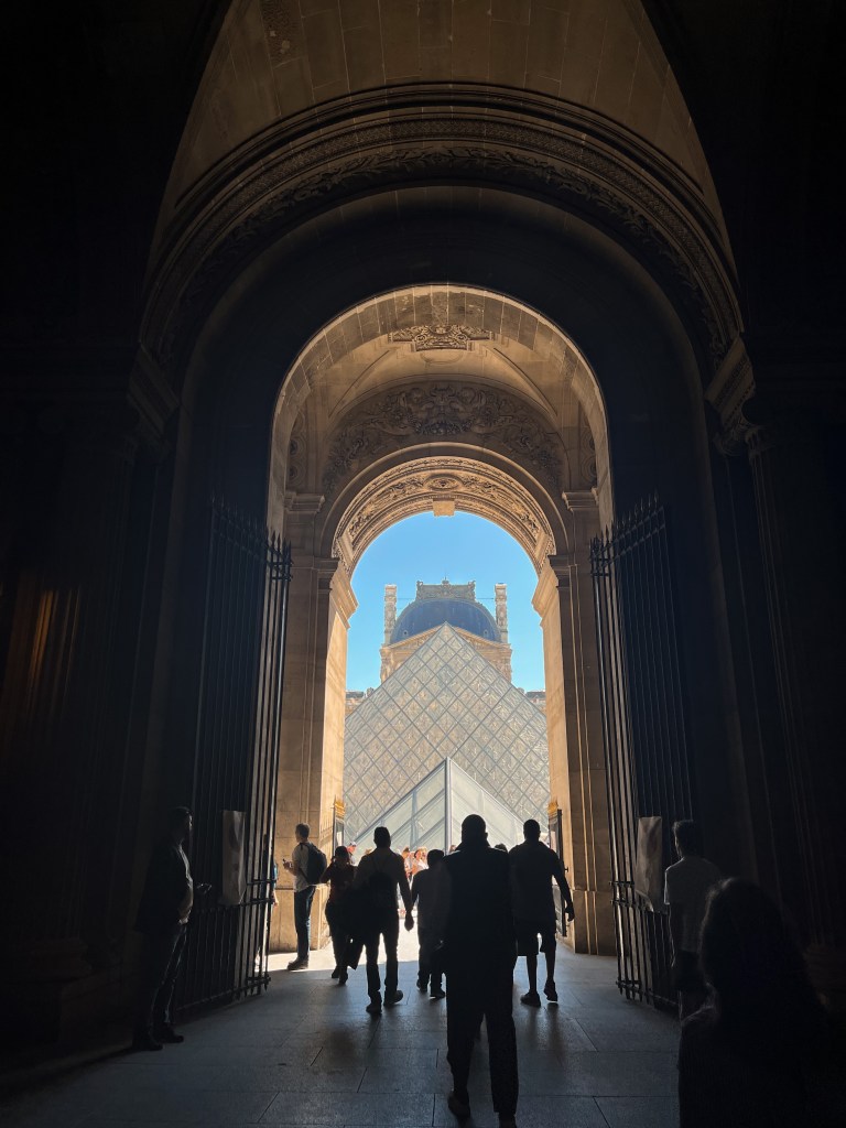 a dark tunnel with light and the glass pyramids of the louvre framed at the end arch. light reflects off tall metal gates