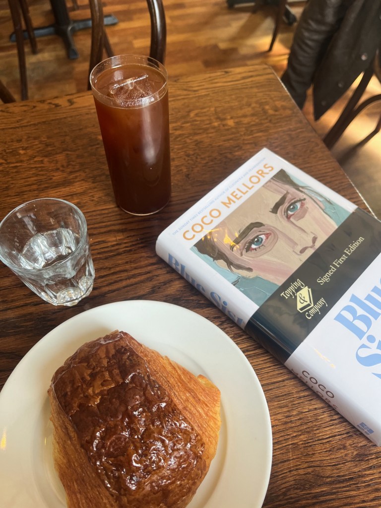 a flat lay on a wooden table with an iced americano, pain au chocolat, hardback book and glass of water.
