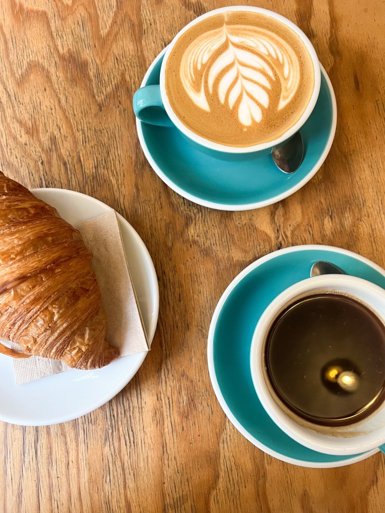 a flat lay style picture of two coffees and a croissant on a wooden table