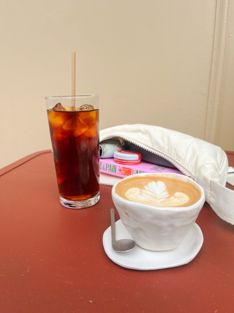an outside table of a coffee shop. There is a flat white, iced black coffee and an open bag laying on the table.