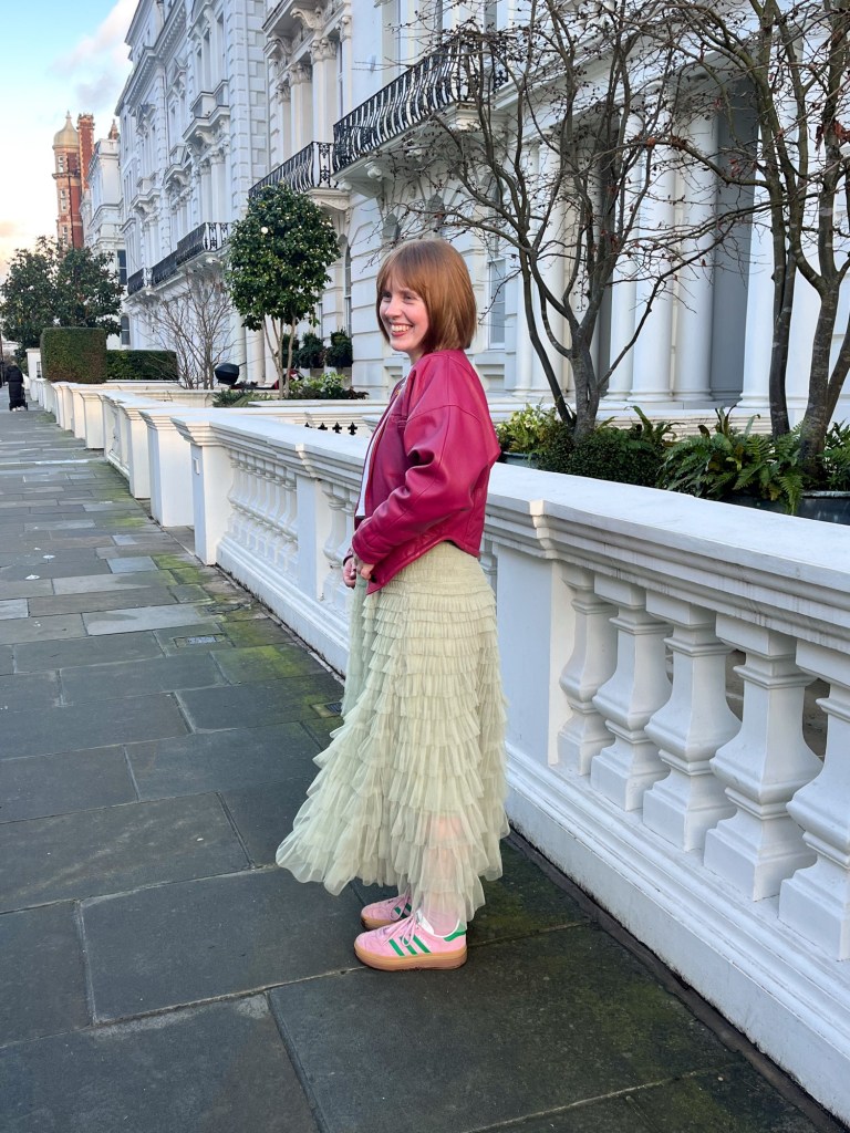 a girl wearing a long, green skirt and pink leather jacket laughing to herself