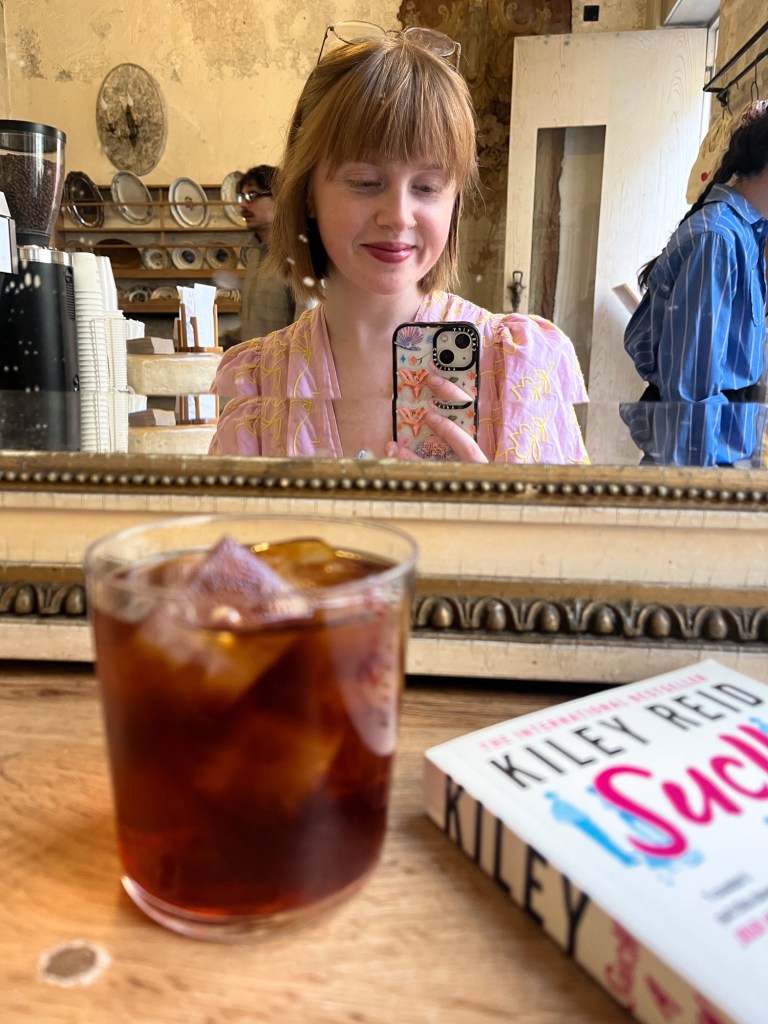 a redheaded girl takes a selfie in a mirror of a coffee shop. There is an iced coffee and book on the table in front of her