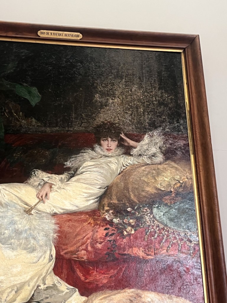 a painting of a woman draped on a red, velvet sofa. she stares straight at you while a dog lies at her feet