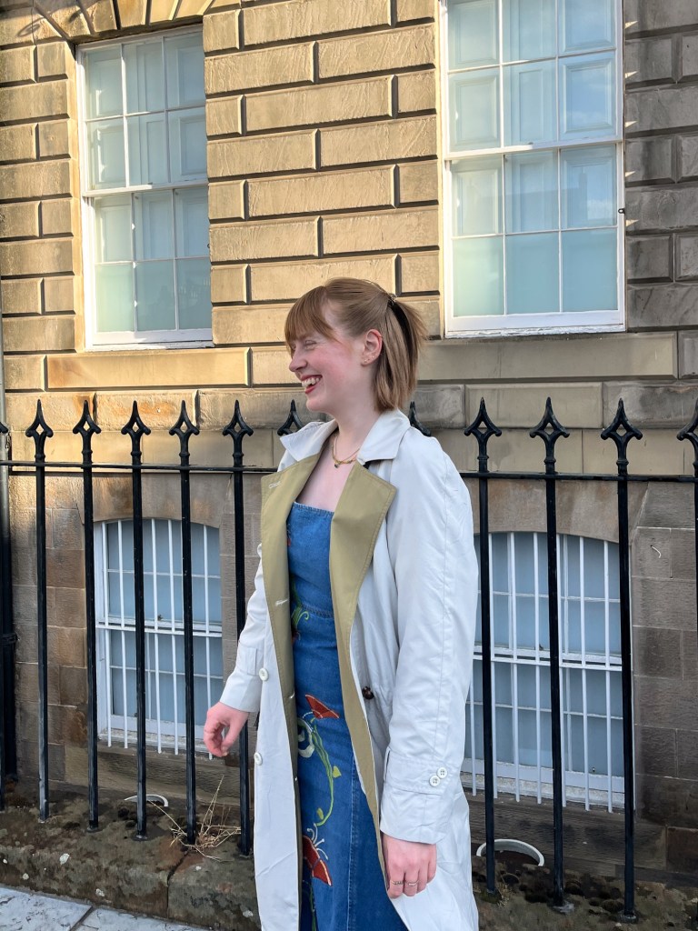 a redheaded girl is laughing. she is wearing an embroidered denim dress and off-white trench coat with green lapels