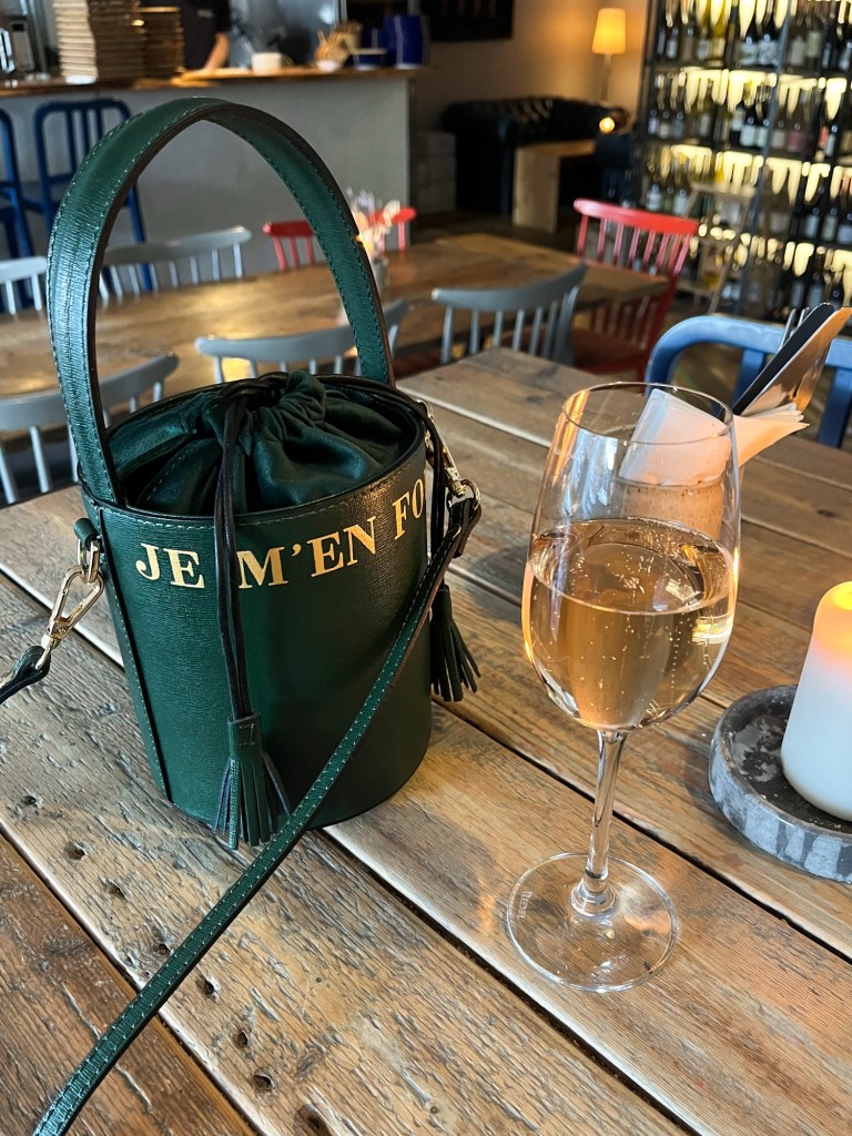 An emerald green bucket bag sat on top of a wooden table. There's a candle and glass of wine sat next to it.