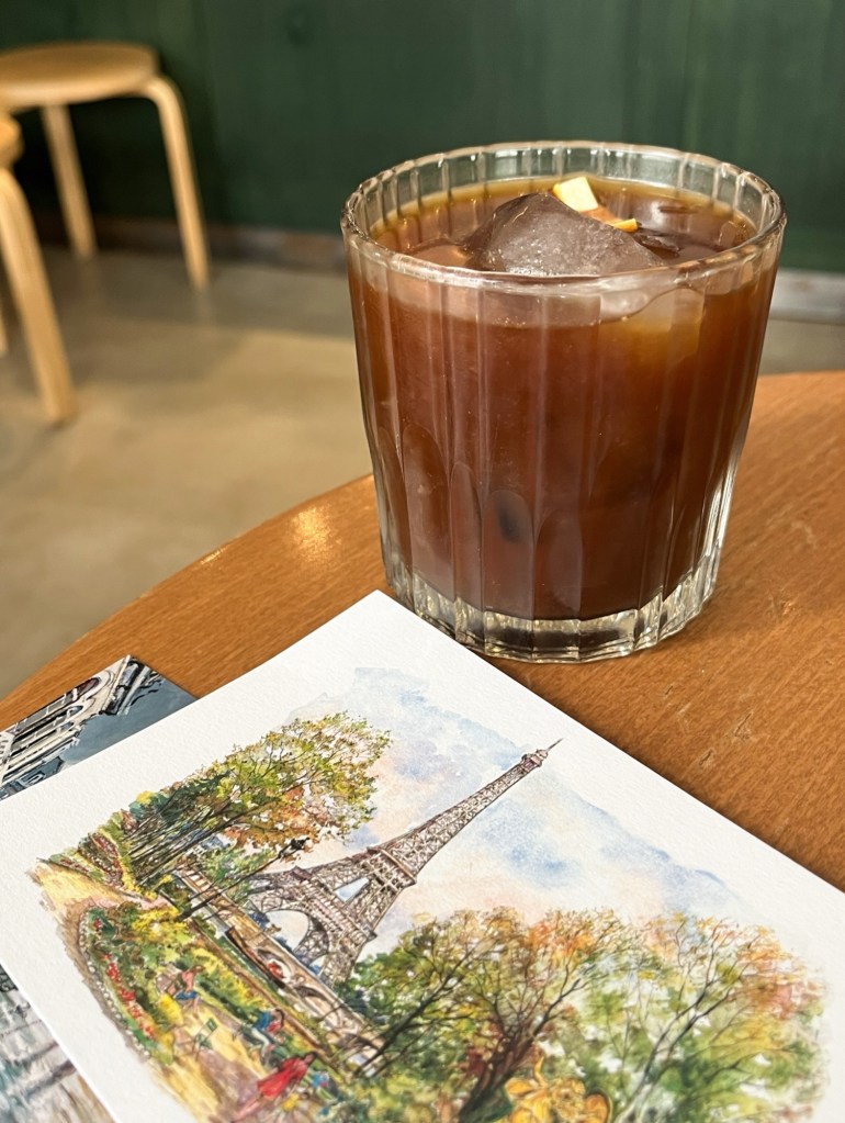 a glass of black cold brew coffee with an orange peel garnish. the coffee is sat on top of a table with illustrated postcards