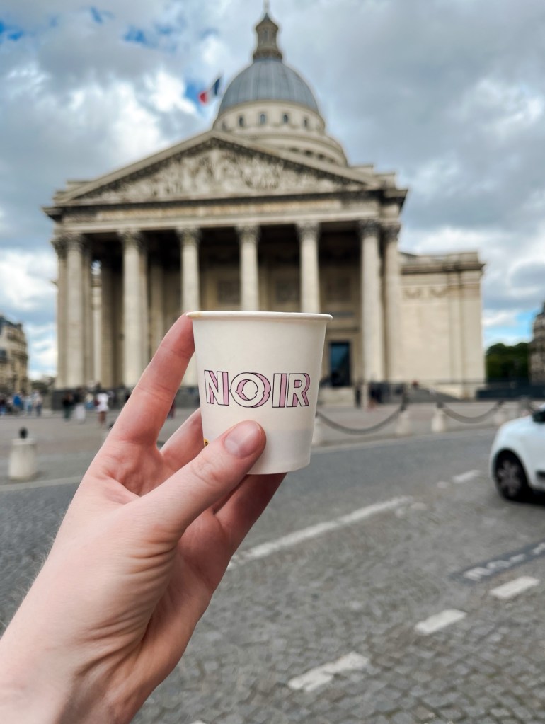 a small espresso takeaway cup is held up in front of Paris's Pantheon