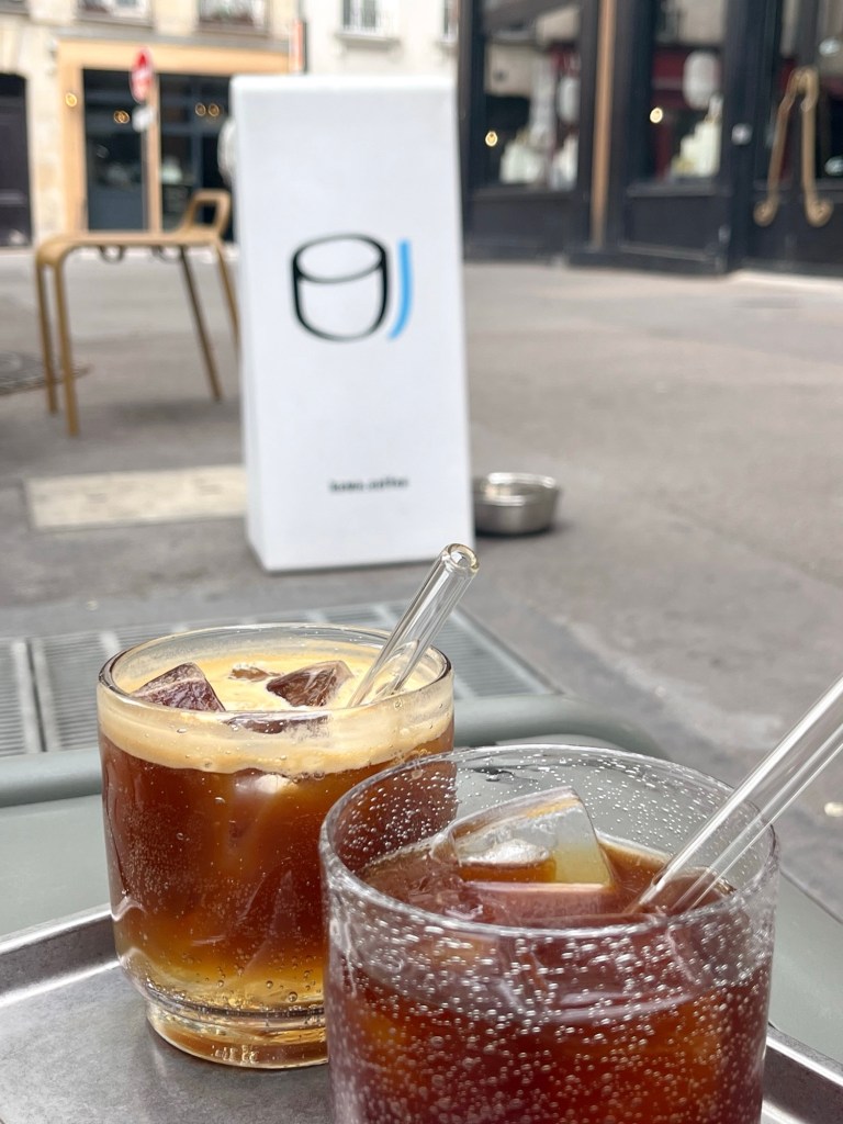Two iced coffees sat on a silver try on the streets of Paris. The coffees are served black with glass straws.