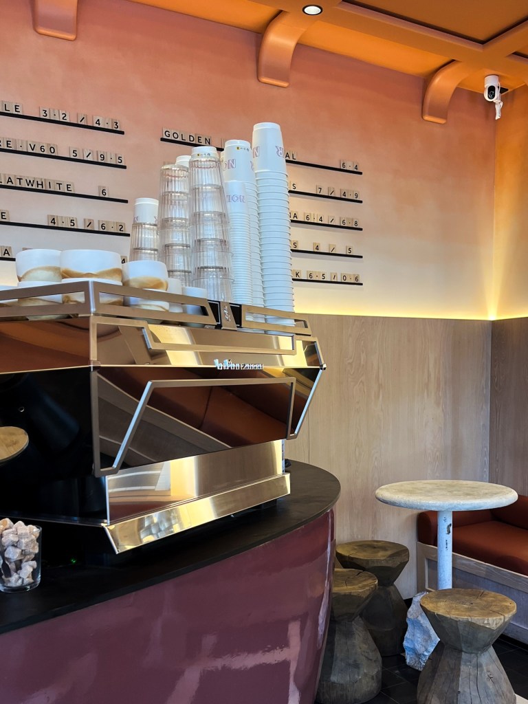 a peachy/purple interior of a coffee shop with warm lighting. There are lots of takeaway cups stacked on top of the coffee machine