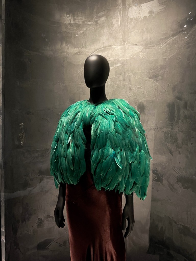 a mannequin wearing a brown silk dress and an emerald green bolero made from feathers
